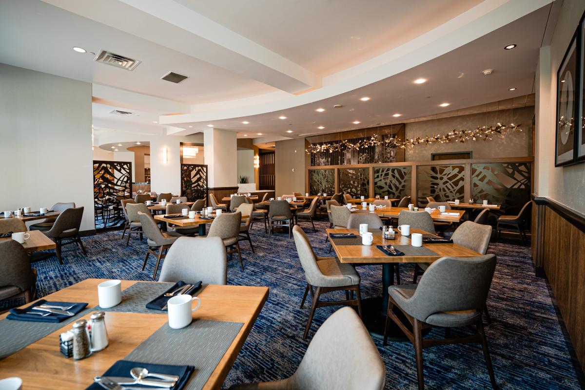 The camera overlooks the new Acqua Restaurant from the back corner of the room. The space features at least twelve tables that each seat 2-4 guests. Each table is set with a modern gray placemat, navy cloth napkins, silverware, plain white coffee mugs, as well as salt, pepper, and sugar. The room dividers are dark solid wood frames with black metal inlays depicting tropical plant fronds. Along the entryway is a string of decorative glass and metal lights.