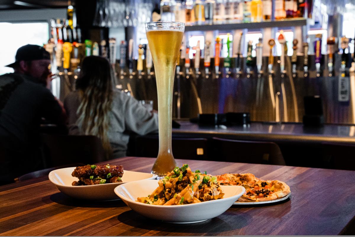 A yard-long beer is featured with three appetizers: a personal-sized pizza, nacho fries, and wings. In the background is a young couple sitting at the colorful Yard House bar.
