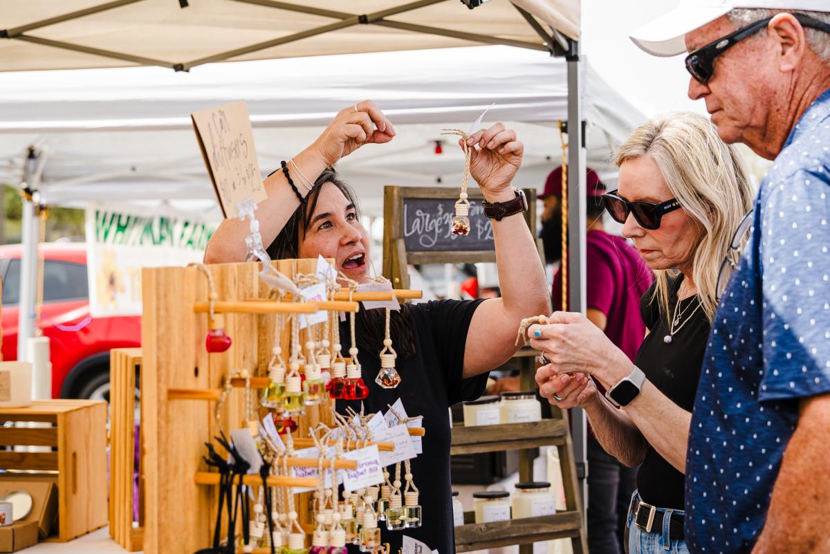 A woman holds up a small glass bottle on a rope chain to an older couple deciding whether to buy. Between them is a display of more bottles in various shapes and containing liquids in different colors.