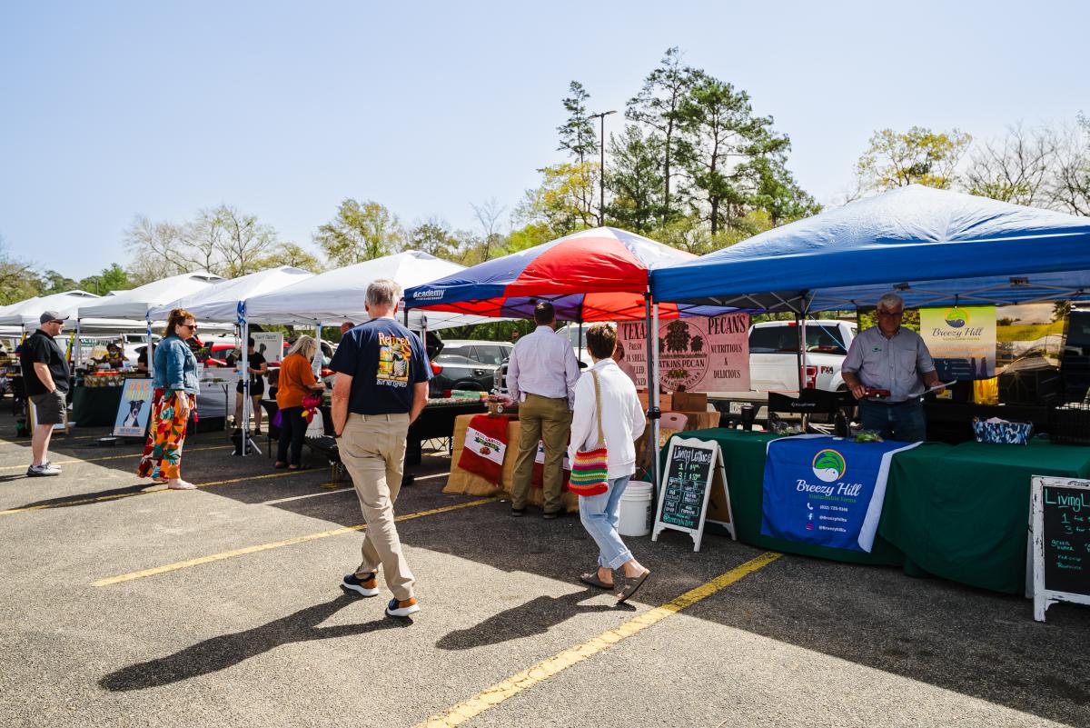 A parking lot is lined with a row of pop-up tents in Texas colors. The visible vendors include "Breezy Hill Sustainable Farms," "Texas Pecans," and "Dog Portraits." Potential customers (men and women of various ages) explore their wares.