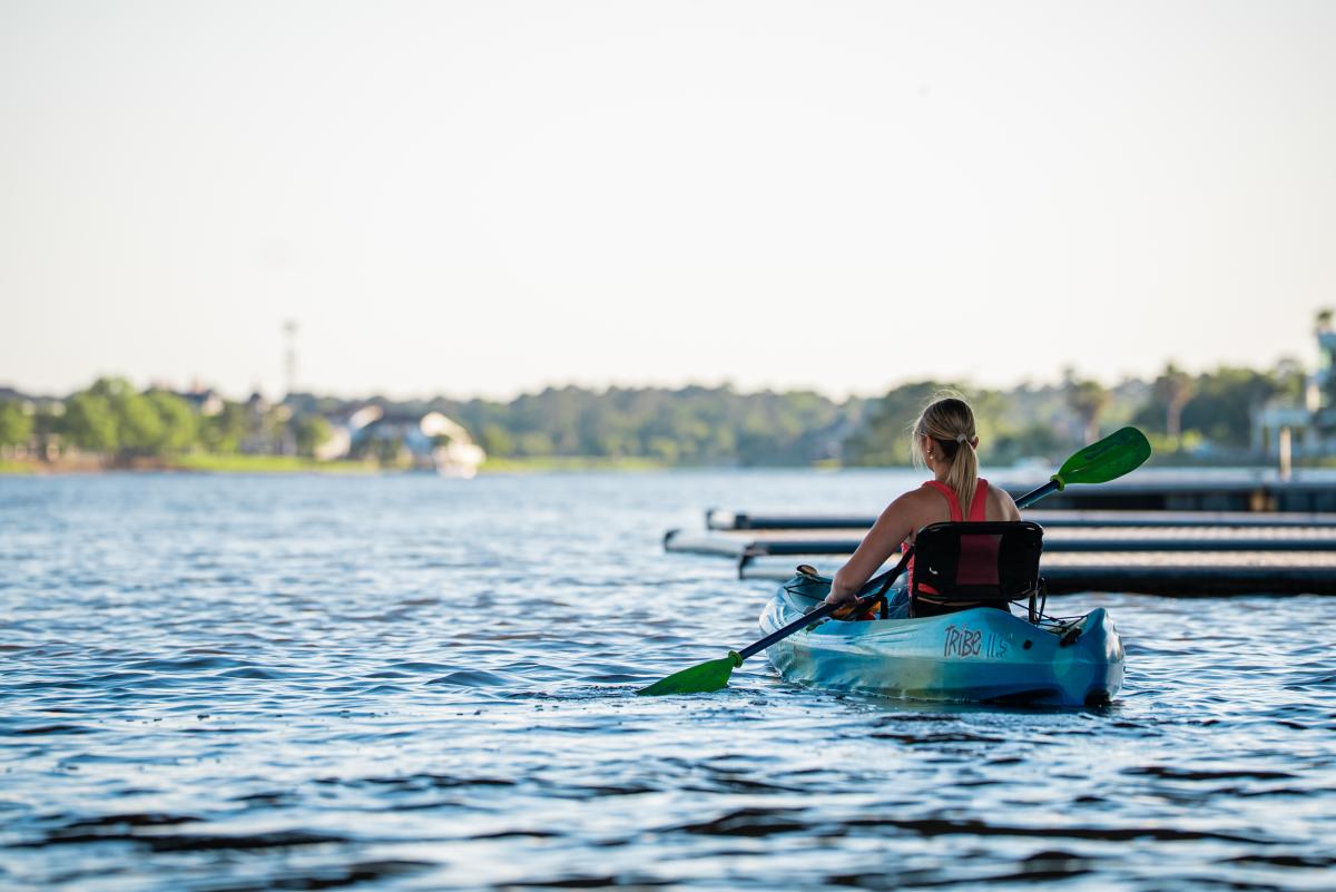 A young blonde woman (Brooklyn Newell) holds a black and green double-sided paddle to row her bright blue kayak. She is facing away from the camera, paddling away from the docks into the open water of Lake Woodlands.
