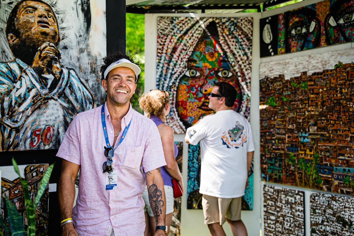 Artist Tony Parana Rodrigues stands in front of his work, smiling at the camera. He appears to be a Latino man in his 30s, wearing a pale pink button-down sport shirt, a white visor, and sunglasses hanging from a WWAF lanyard. He also has a large abstract tattoo on his left forearm. His paintings are colorful, abstract portrait. One is of a young Latino boy praying, his eyes looking to the heavens. Another is of a young girl staring directly at the viewer, her face and headscarf pieced together like a colorful floral quilt. A few others look to be distant skylines of other parts of the world.