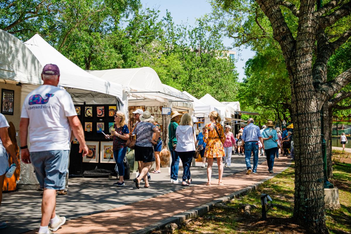 White pop-up tents line The Waterway, where dozens of visitors are seen admiring the artwork on display. The walkway is shaded by thick green trees.