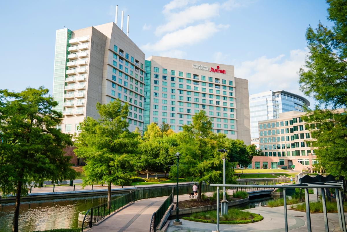 The exterior of The Woodlands Waterway Marriott from the view of someone walking along a stretch of The Waterway behind the hotel. The sky is light blue with small white clouds drifting overhead. The trees along the water are thick and bright green.
