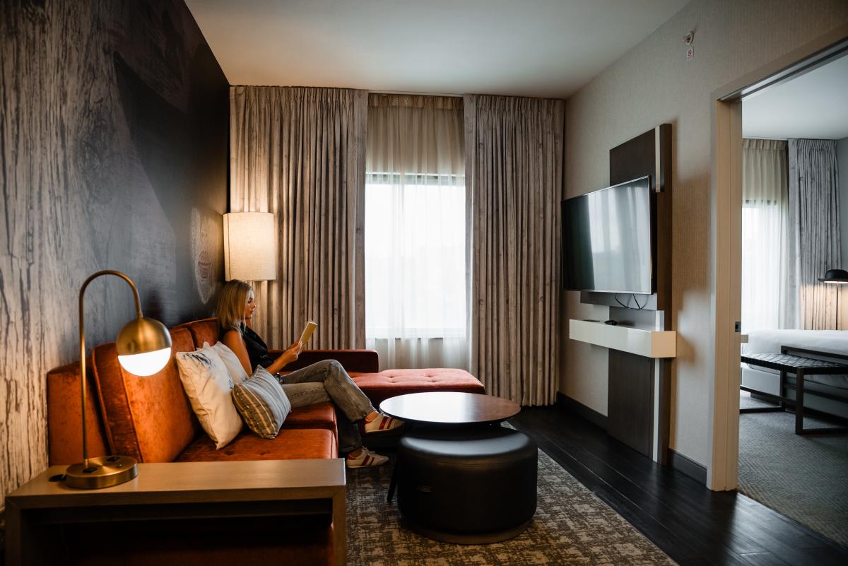 The common area of a Hyatt Centric King Suite features a long red velvet couch along the left wall, facing a flat-screen TV. There is a small, round coffee table and an ottoman that fits underneath. Beside the couch, closest to the camera, is a round copper lamp. The curtains along the far wall are half open, filling the space with light. A young blonde woman in a black blouse and blue jeans sits in the center of the couch, reading a book. To the right is a door to the bedroom, where part of a king bed is visible.
