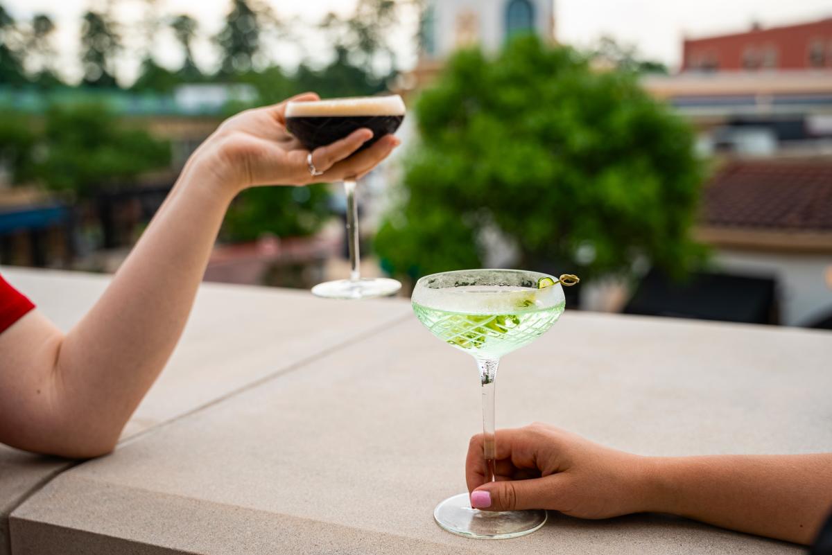 Two women (out of frame except for one arm each) enjoy martinis from Hyatt Centric Kitchen & Bar on the Hyatt Centric terrace. On the left, a woman rests her elbow on the balcony overlooking Market Street with an espresso martini cupped in her upright palm. On the right, her companion rests her arm along the balcony, her fingers wrapped loosely around the base of a crisp green cucumber martini.