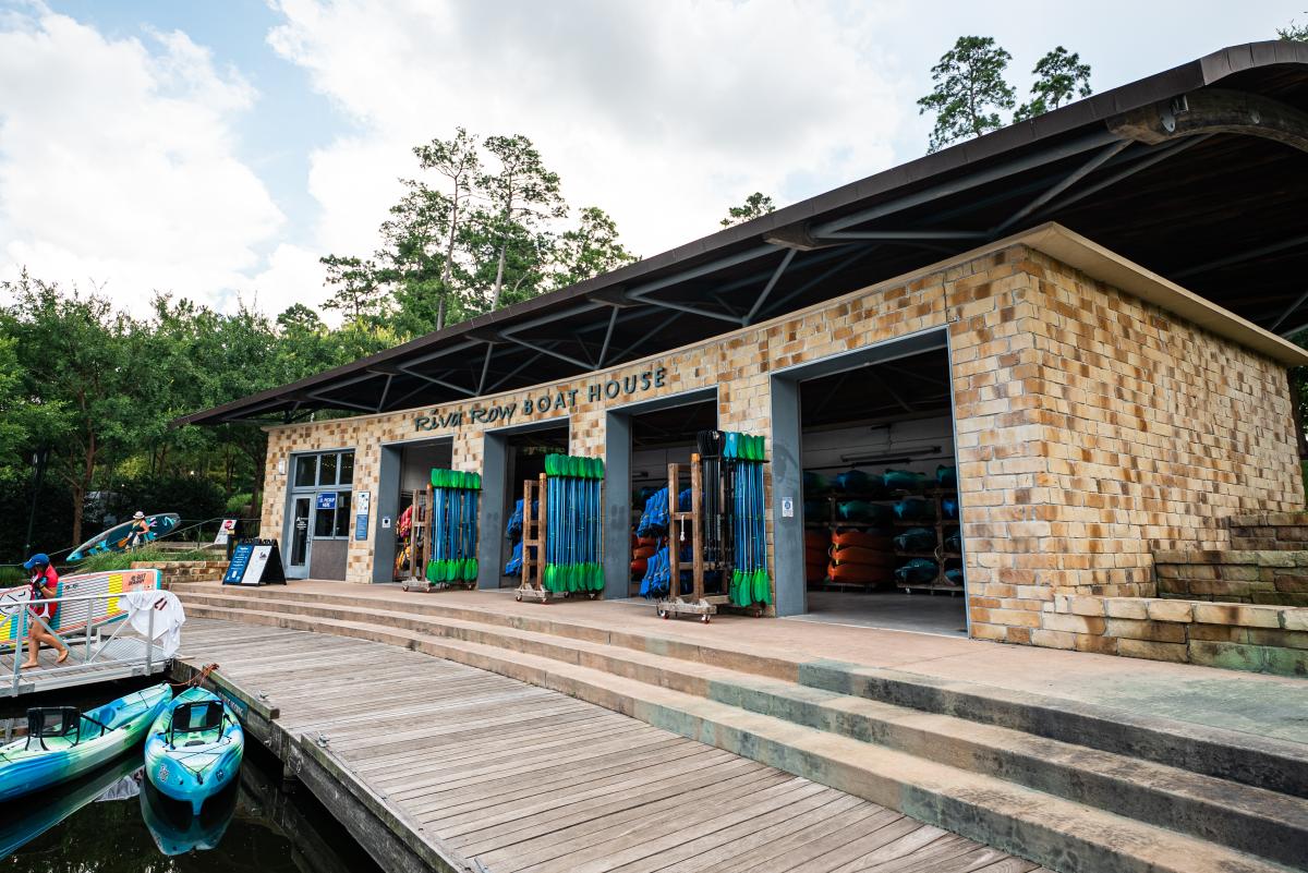 The boat house is a long brick building with four open garage-like doors and a registration office on the far end. Inside are shelves stacked with kayaks in various colors. Outside the doors are standees with dozens of double-ended paddles. The building sits on a dock, where two kayaks rest on the water.