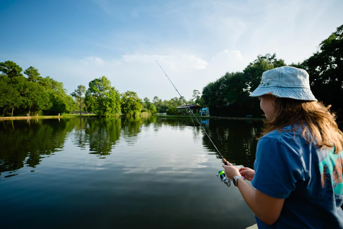 A young woman (on the right of the frame) stands on the shore of the stock pond at Creekwood Park. She is wearing a denim-blue shirt with sea turtles on the back as well as a denim bucket hat with white flowers embroidered around the front. She is holding a fishing pole made of wood and green plastic. The water is dark and gently rippling. In the background is a covered pavilion for picnics, bright green trees, and a pathway toward (in the distance) tennis courts.