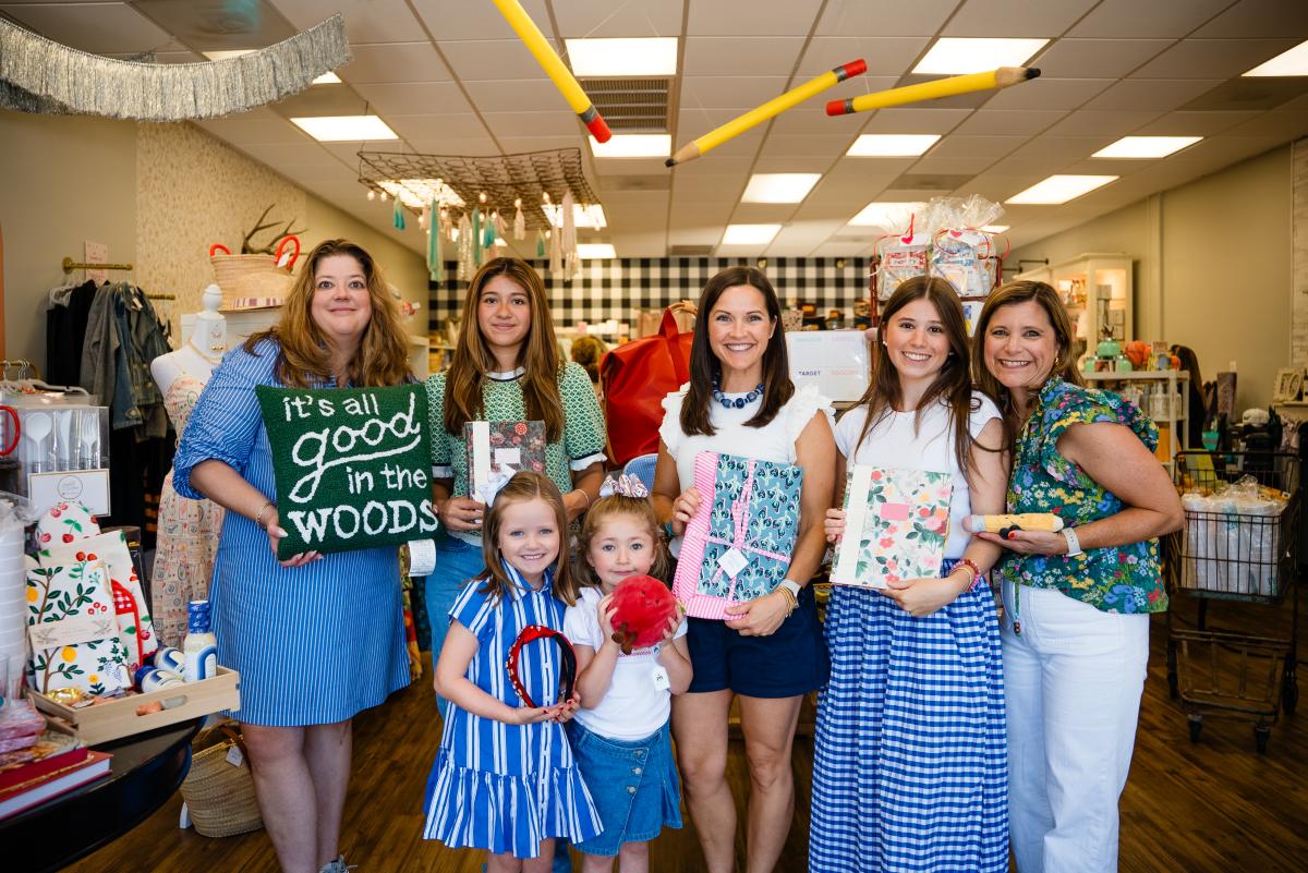 A group of seven women/girls smile and present boutique gifts to the camera. They are inside Tumbleweeds & Notions, a local gift shop, promoting cute back-to-school shopping supplies. One woman holds a fuzzy green pillow that reads, "it's all good in the woods." Two teen girls hold floral notebooks. Two young girls hold up a sparkly red headband and a red apple plush.