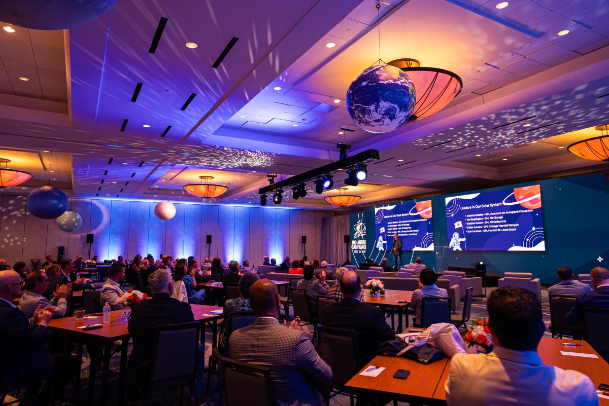 The viewer is in the audience of the Marriott General Managers Meeting at The Woodlands Waterway Marriott. There is a speaker on stage with a PowerPoint presentation listing GMs receiving an award. The room is lit up with deep blue, purple, and orange lights to simulate outer space. Around the room are various inflatable planets hanging from the ceiling.