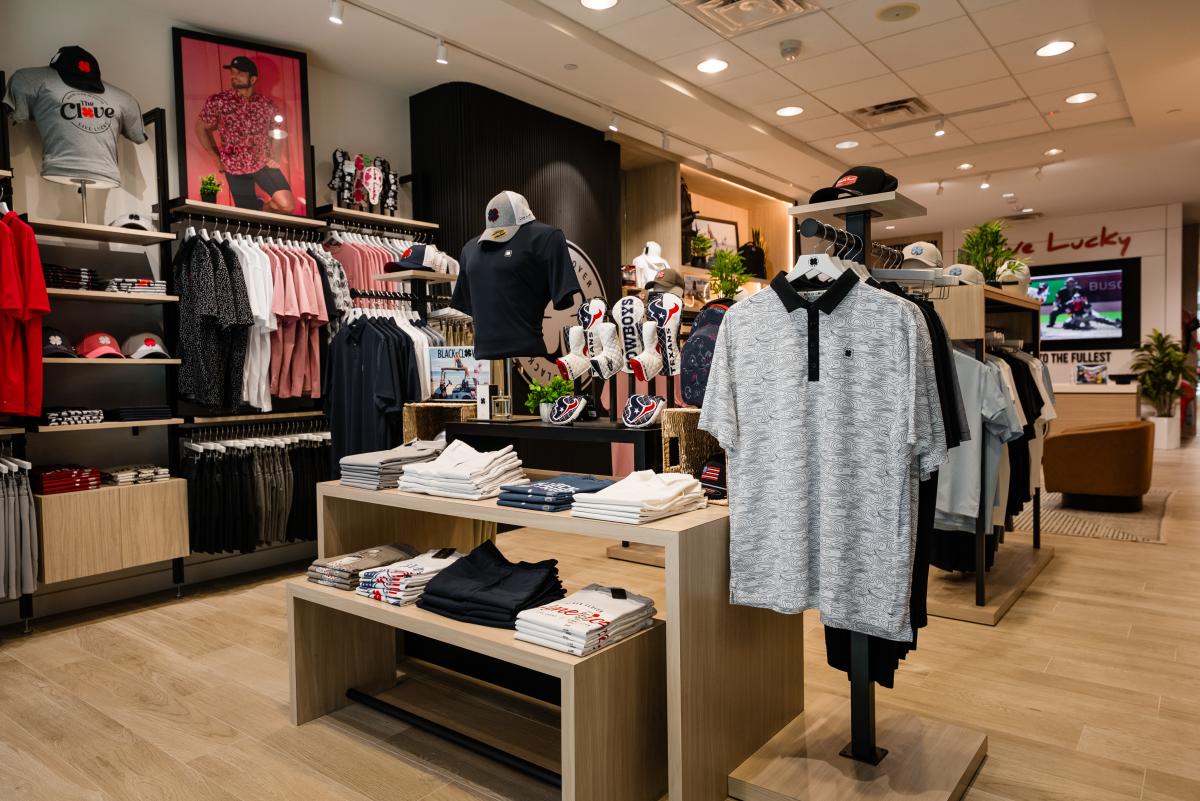 The camera overlooks the interior of the Black Clover at The Woodlands Mall. The central display and the wall on the right feature both folded and hanging clothes: sport shirts, slacks, ballcaps, and golf club covers.