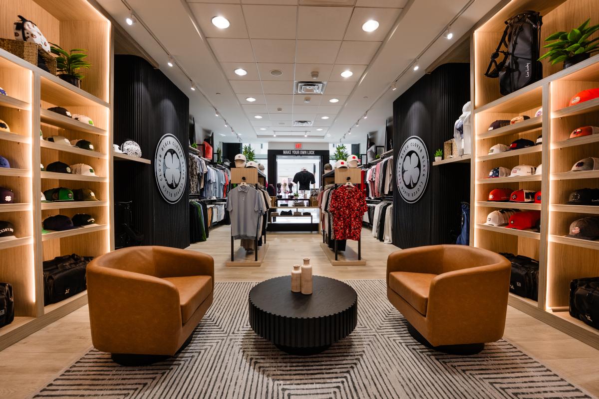 The camera overlooks the interior of the Black Clover at The Woodlands Mall. Closest to the camera is a small sitting area with two brown leather chairs and a black, round coffee table. Behind it are three displays of various sporting apparel. Along the side walls are wooden shelves lined with hats and large logos of a white clover on a black background.