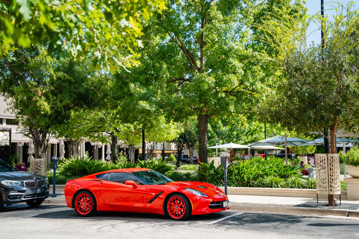 A bright red sports car is parked along the sidewalk at Market Street. The trees are bright green. People relax under patio umbrellas in the background.