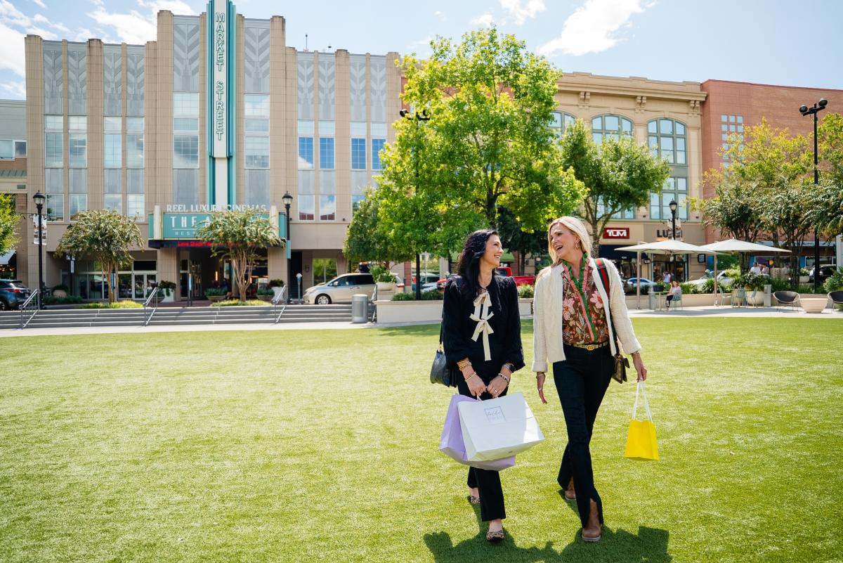 Two women dressed for fall walk through Central Park at Market Street. The blonde woman (right) is carrying a small yellow shopping bag from Kendra Scott. The dark-haired woman (left) is carrying larger bags in purple (Hemline) and white (Golden Gray Boutique).