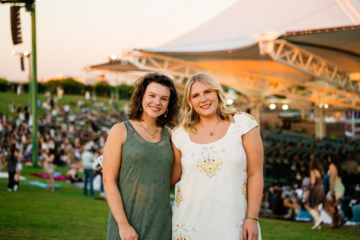 Two young women smile at the viewer during a show at The Cynthia Woods Mitchell Pavilion. The girl on the left has dark curly hair and is wearing a casual olive green dress. The girl on the right has blonde hair and a white dress embroidered with colorful flowers.