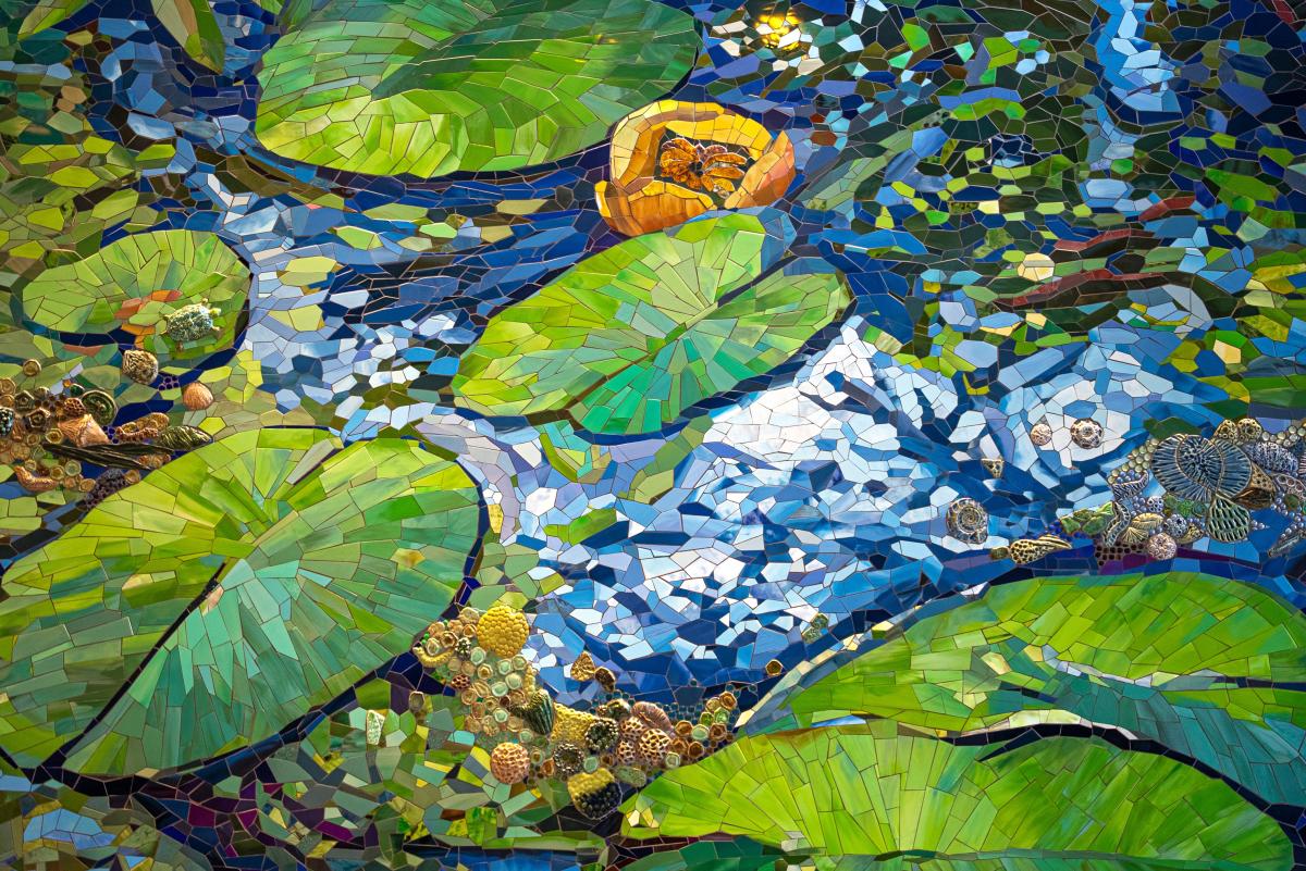 Reflections to Mist Mosaic in Stairwell at Sam Houston State University - The Woodlands. The full art piece is 4 stories tall, wrapping around the interior and exterior of the building. This segment (ground floor) is a close-up of bright blue water, bright green lily pads, bright yellow flowers, and 3D coral- or fossil-like accents.