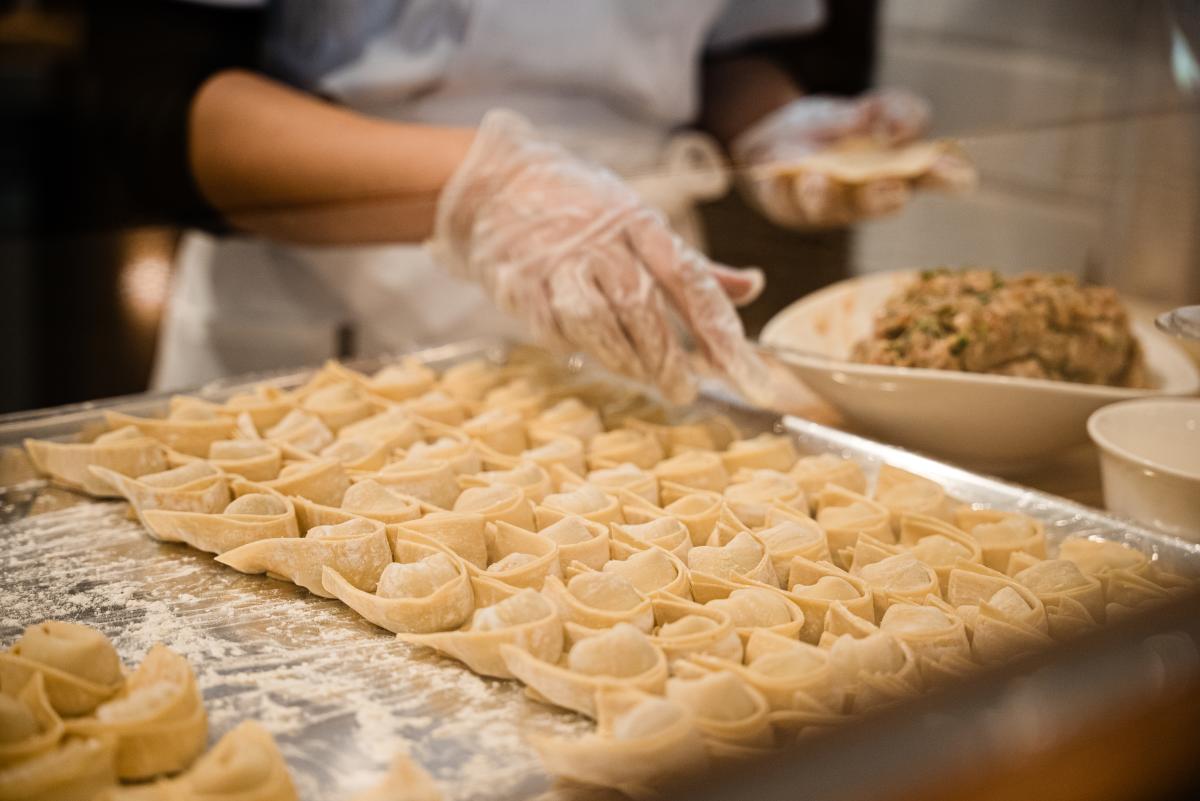 The camera focuses on a metal baking tray of neatly folded dumplings. Behind it are the hands of the chef, one holding a small circle of dough, the other reaching for a bowl of the stuffing.