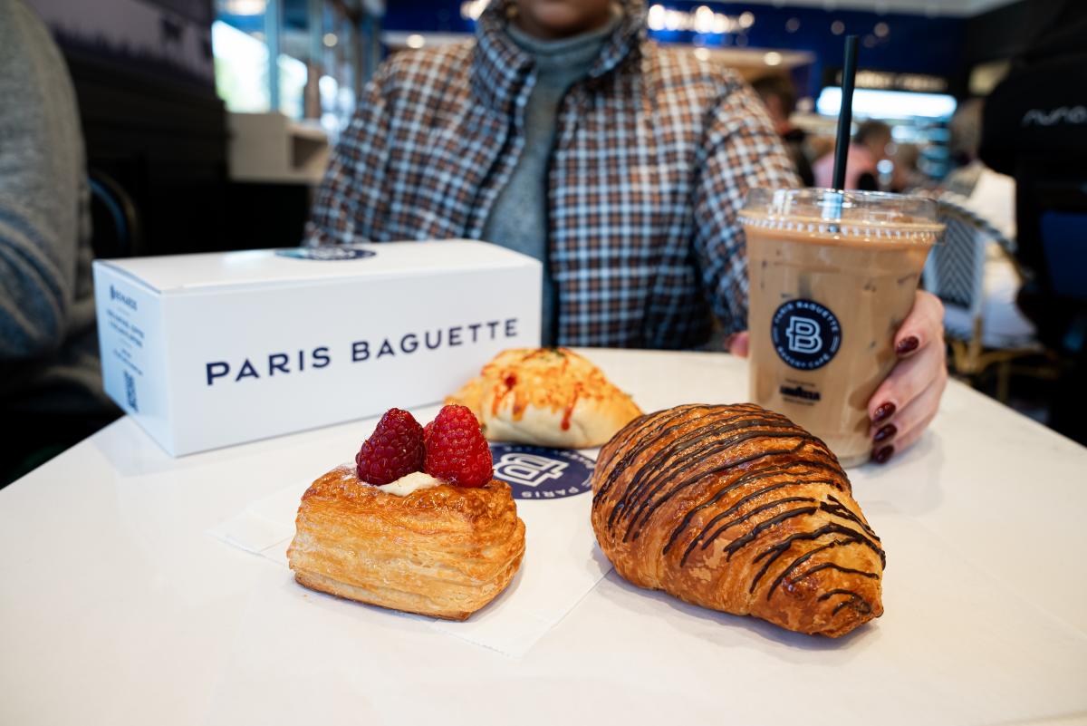 The camera focuses on a table set with pastries (a puff pastry topped with raspberries and cream, a chocolate-drizzled croissant, and a melted cheese biscuit). Behind it is the box it came in, the "Paris Baguette" name angled neatly for the camera. A woman sitting behind the table--her head out of the frame--holds a cup of iced coffee in a Paris Baguette to-go cup.