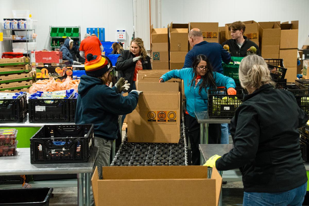The VTW Sales & Marketing Committees volunteer at the MoCo Food Bank. The camera looks down the length of a conveyor belt. Five women and two men stand on either side of the belt, counting various types of produce and placing them in boxes for shipping.