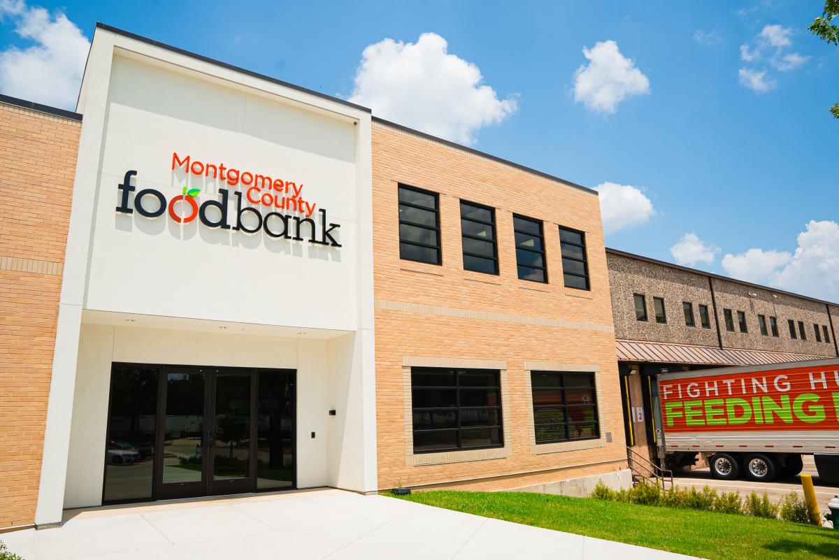 The MoCo Food Bank is a tan-brick building with lots of windows and a white entrance with the full name of the organization in bright red and black text. The second "o" in "food" looks like a minimalist apple. On the right is a docked 18-wheeler that reads, "Fighting Hunger | Feeding Hope."