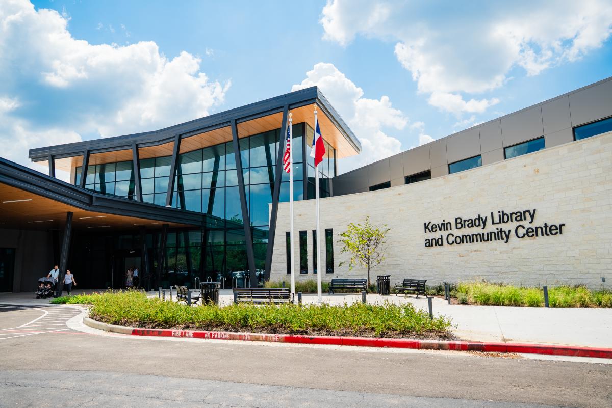 The Kevin Brady Library is a long, angular building. The section on the right is made of pale brick with the name in large black letters. The section of the left is taller with floor-to-ceiling windows and thin black pillars. The roof makes an ever-so-slight "V" shape, revealing a bright copper-colored ceiling underneath. The entrance is shaded by an awning matching the roof. Two flagpoles stand tall in the middle of the frame.