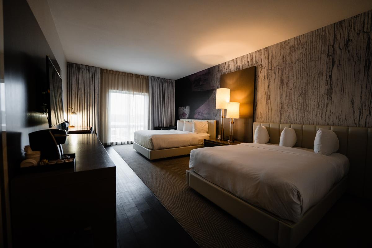 The Hyatt Centric Double Queen Suite features two queen beds along the right wall with a nightstand and two large lamps in between. There is a large dresser and writing desk on the opposite wall and a long window with thick curtains along the back wall.