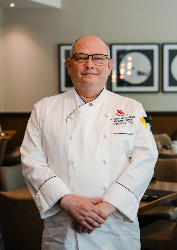 A man, 40s, smiles at the camera in The Waterway Marriott's Acqua Restaurant. He's wearing glasses and a white chef's uniform that reads, "MARRIOTT | Woodlands Marriot | Michael Batke | Executive Chef" over his left breast pocket. He is clasping his hands in front of him. Behind him are a few square breakfast tables and framed abstract art on the wall.