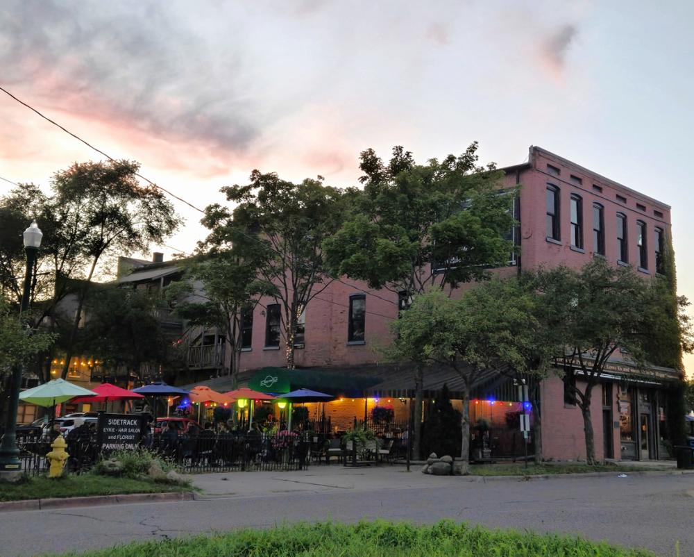 Evening setting, view of back side of Sidetrack Bar and Grill with patio seating, colorful umbrellas, and lighting illuminating the patio area. Green trees and sunset skies fill the photo.
