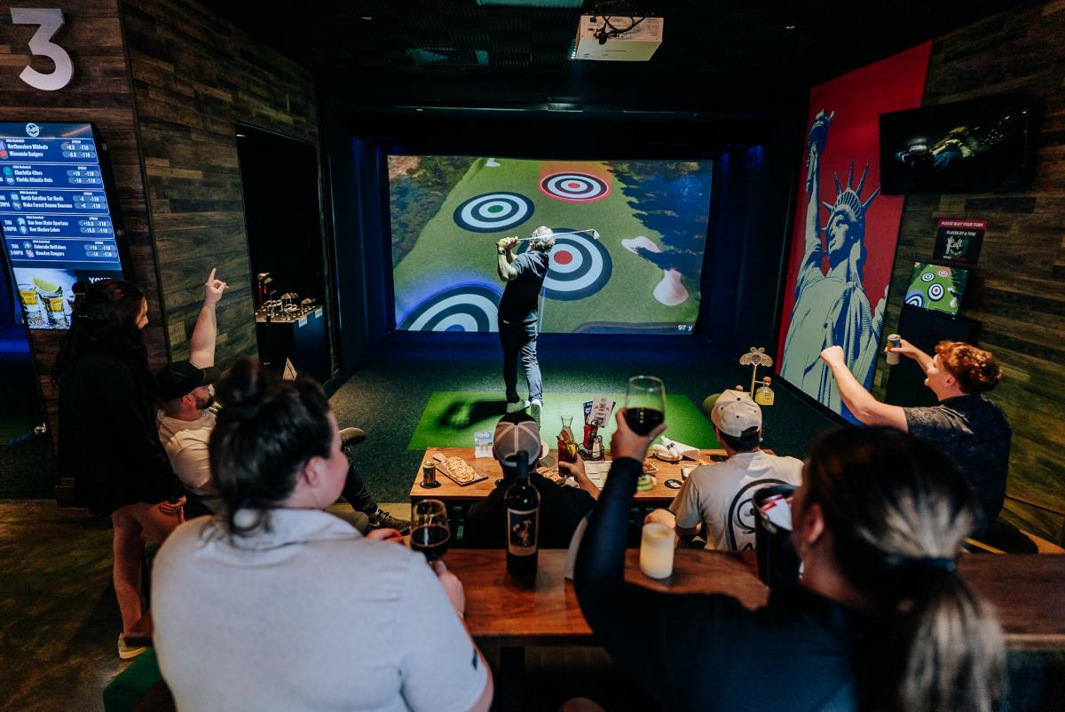 A group of people cheer on a man practicing his golf swing with virtual golf.
