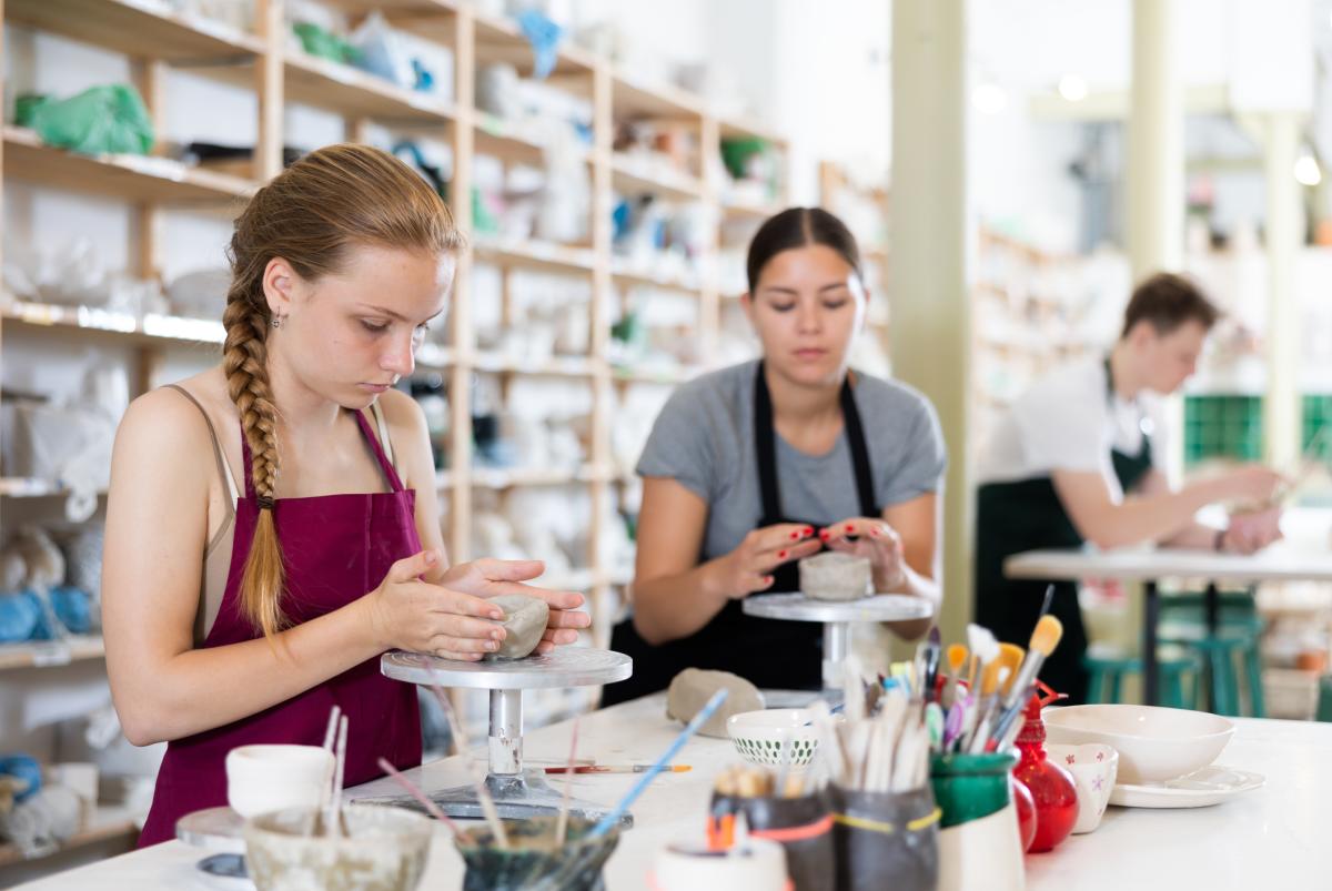 Two teenage girls sit at a table with pottery, paints, and paint brushes in a pottery studio.