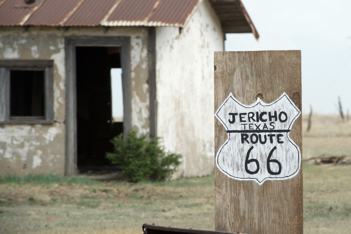 A hand-painted Route 66 sign marks Jericho, Texas in front of a weathered adobe building with peeling paint and a rusty tin roof. Set against the windswept plains, this quiet stop offers a glimpse into the ghost towns that once thrived along the historic Mother Road.