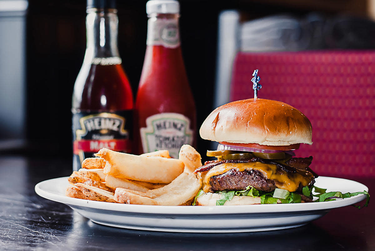Cheeseburger with melted cheese, bacon, lettuce, red onion, and pickles on a bun, served with potato chips on a white plate, with condiment bottles in the background.