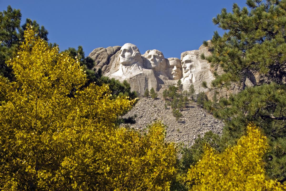 Mount Rushmore with four presidential faces carved into the granite, backed by a clear blue sky. Vibrant yellow autumn foliage in the foreground.