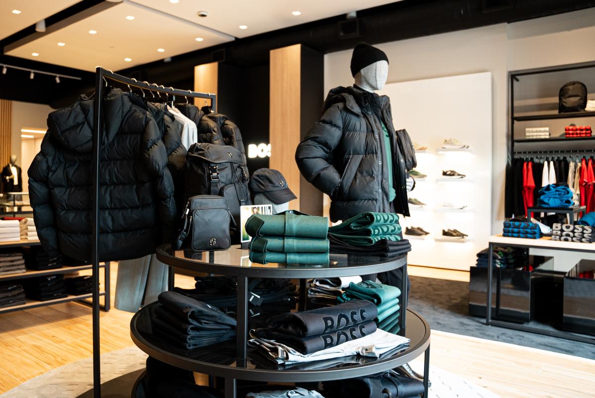 The interior of a BOSS Menswear store, facing the main display. A round three-tiered table is topped with folded athleisure shirts, bags, and hats, most of which are black or bright green. A coatrack behind the table features black and gray puffy jackets. A mannequin models these latest fashion piece.