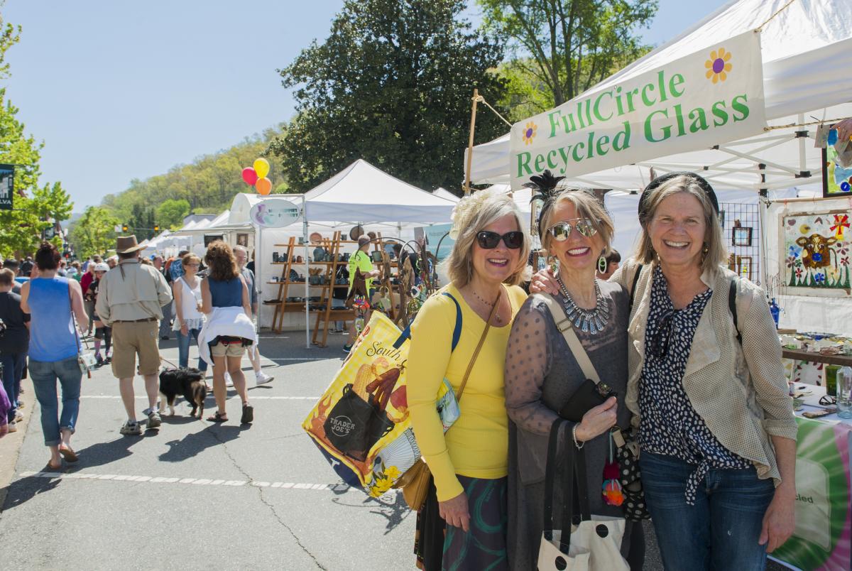 Three women smile for a photo at an outdoor street festival. They are standing in front of a white vendor tent labeled "FullCircle Recycled Glass" on a sunny day with many other festival-goers and tents in the background.