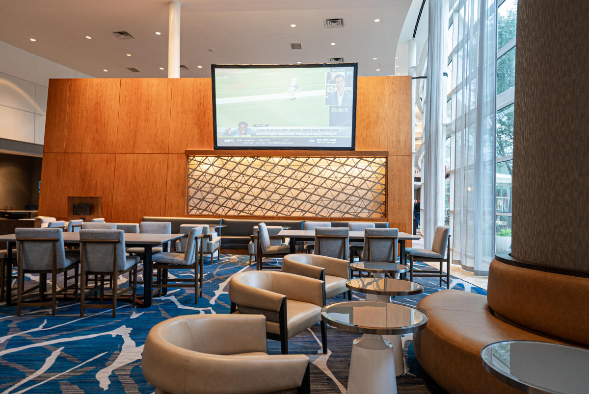 The camera faces the lobby sitting area at The Woodlands Waterway Marriott. In the foreground are three small tan cushioned chairs, each with their own miniature table, circling a decorative support pillar of the building. Further back are two large breakfast tables that seat six people each. A dividing wall in the very back features decorative latticing and a giant flat screen TV. To the right are floor-to-ceiling windows that illuminate the room.