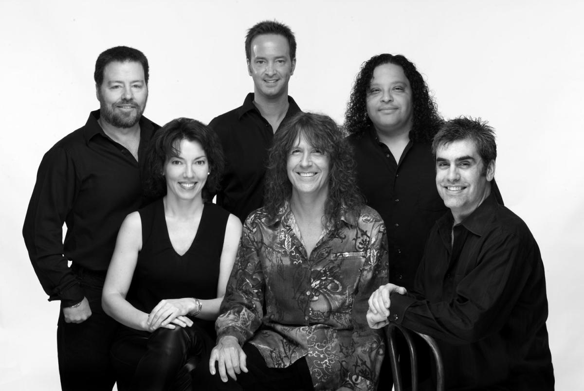 A black-and-white portrait of Windborne Music's Led Zeppelin Group. There are five men and one woman (front row, left of the frame). They are all dressed in black button-down shirts except for the woman (a black blouse) and the man in the front/center (a lighter, likely-brown shirt with a black and white vine-like pattern.