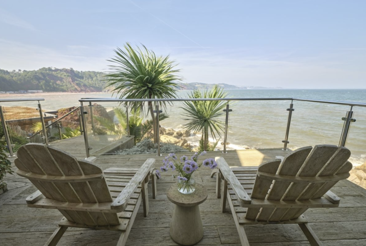 Two wooden lounge chairs on a deck overlook a serene ocean view, framed by tropical plants and a glass railing, with a vase of purple flowers on a small table between them.
