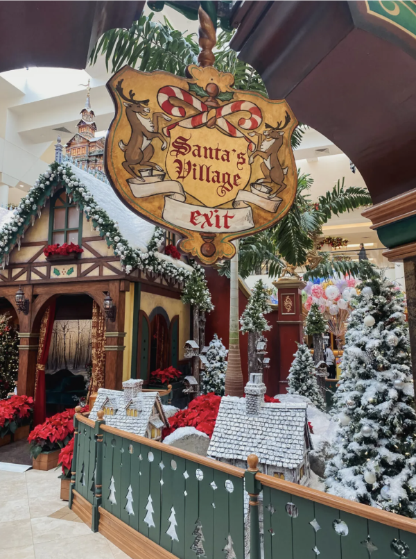 Image a house covered in snow and decorated with Christmas decor. In the top middle section of the picture, a wooden sign can be seen hanging. It's yellow with red and white text. It reads "Santa's Village." Snow covered trees can be seen in the background next to the house.