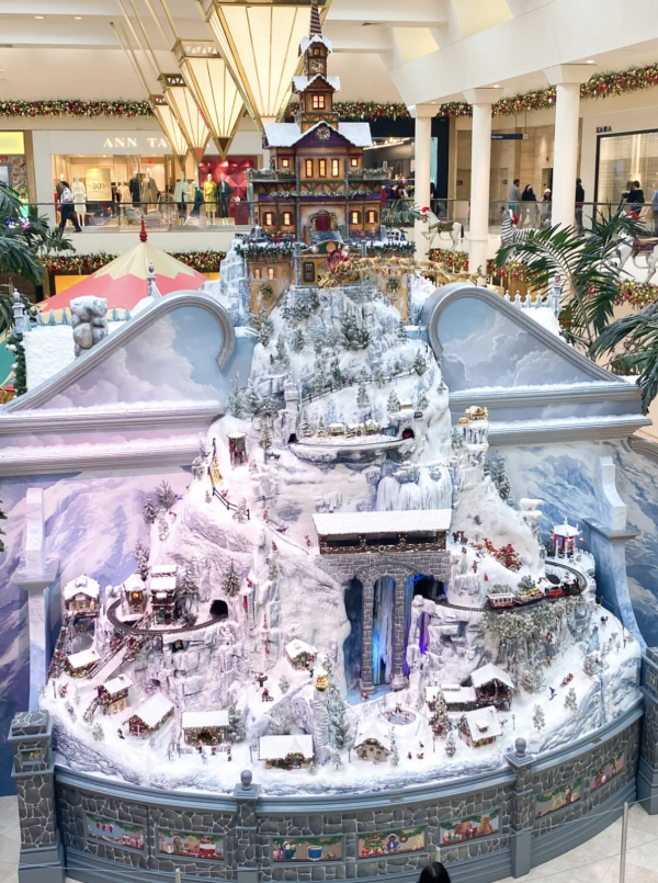 Image of Christmas decorations set up in the middle of South Coast Plaza. The decorations include a snow covered village, with trees and a train.