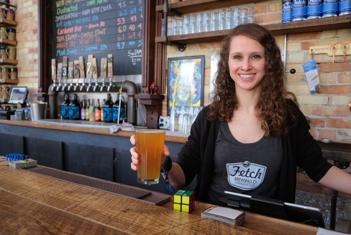 smiling woman with long curly brown hair holds pint glass of craft beer up from behind bar of fetch brewing company