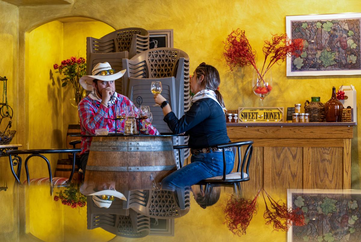 In a warmly lit room with yellow walls, two individuals sit at a table made from a barrel, sharing drinks. The room is decorated with a sign reading "Sweet & Honey," a colorful map, and vases filled with red twig branches.