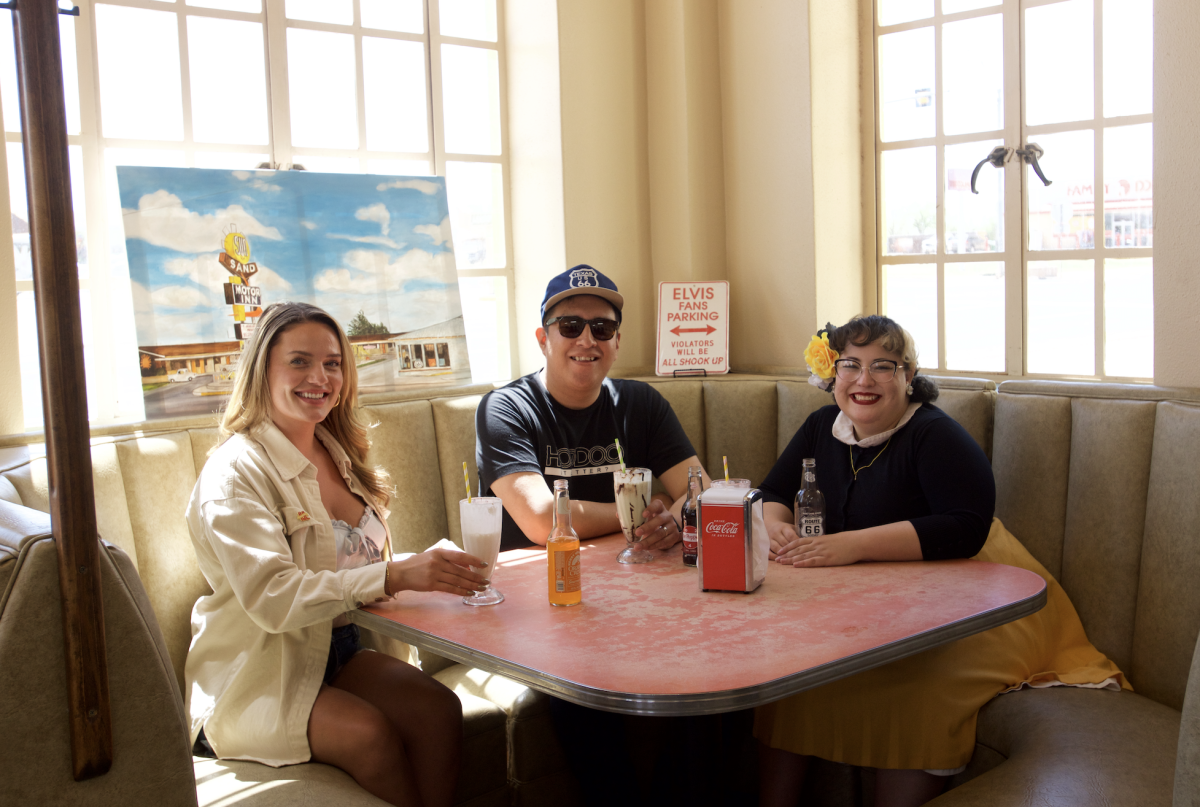 Three friends enjoy milkshakes and bottled sodas in a retro diner booth filled with sunlight, surrounded by vintage decor. A playful “Elvis Fans Parking” sign and a painting of the iconic Sands Motel add nostalgic Route 66 charm to the cozy, Americana vibe.