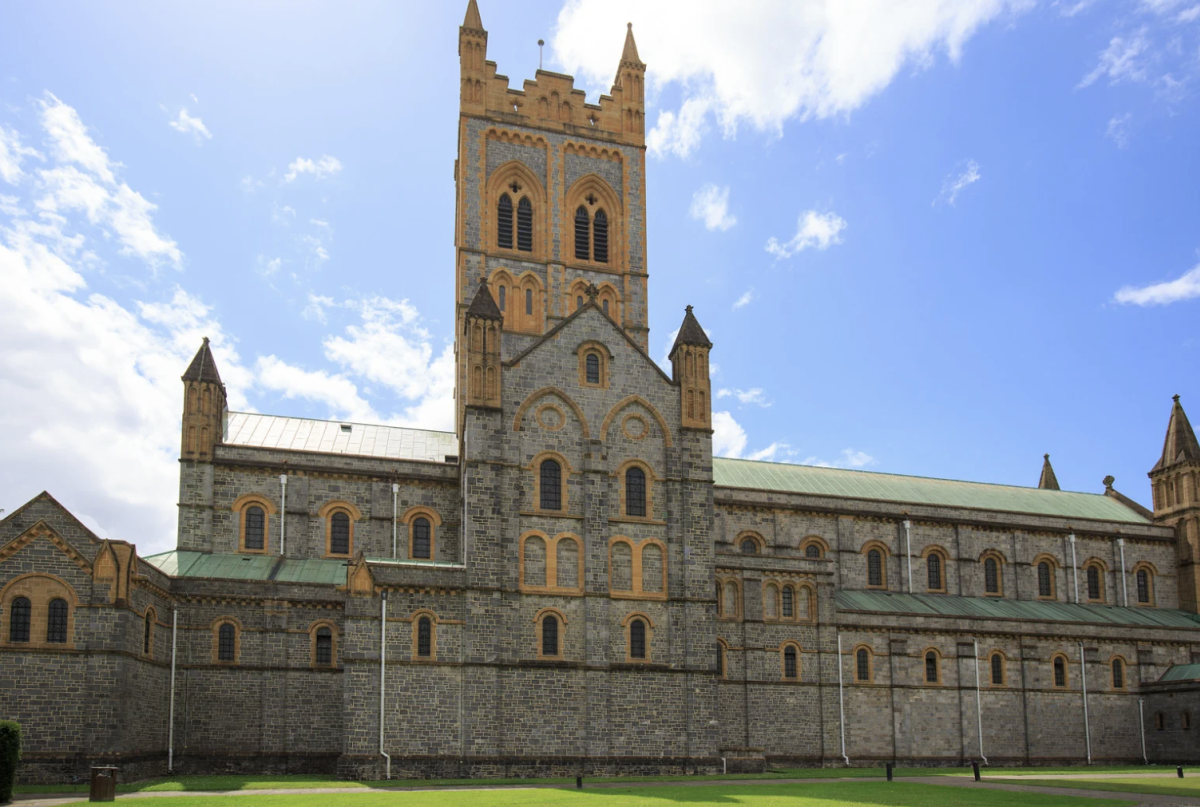 A large historic cathedral with a central tower and pointed arches, set against a bright blue sky with scattered clouds. The tone is majestic and serene.