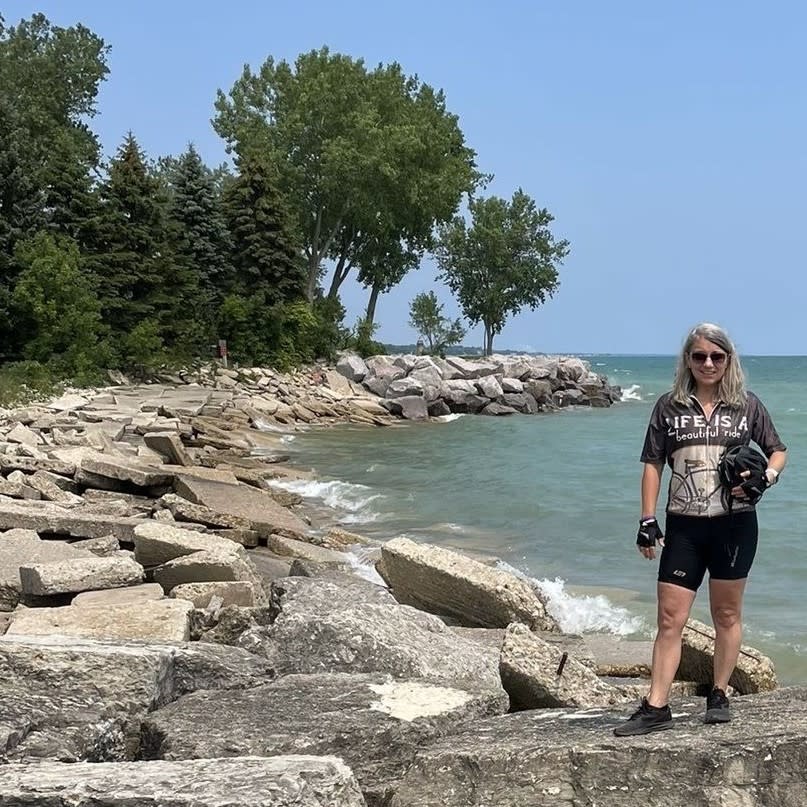 woman in cycling gear standing on rocks by Lake Michigan with trees and blue sky in the background