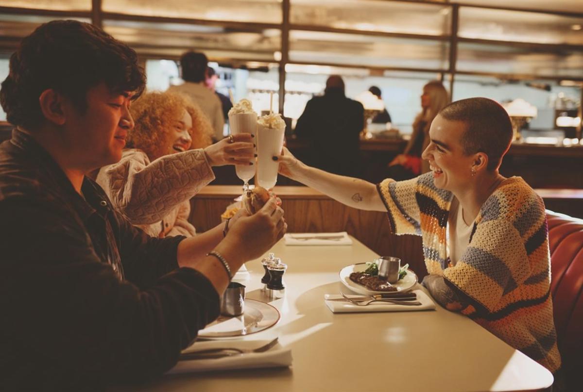 Two people seated in a booth at a diner, raising tall milkshakes towards each other across a table. Desserts and drinks are arranged on the table, while warm lighting and other diners create a cosy, relaxed atmosphere in the background.