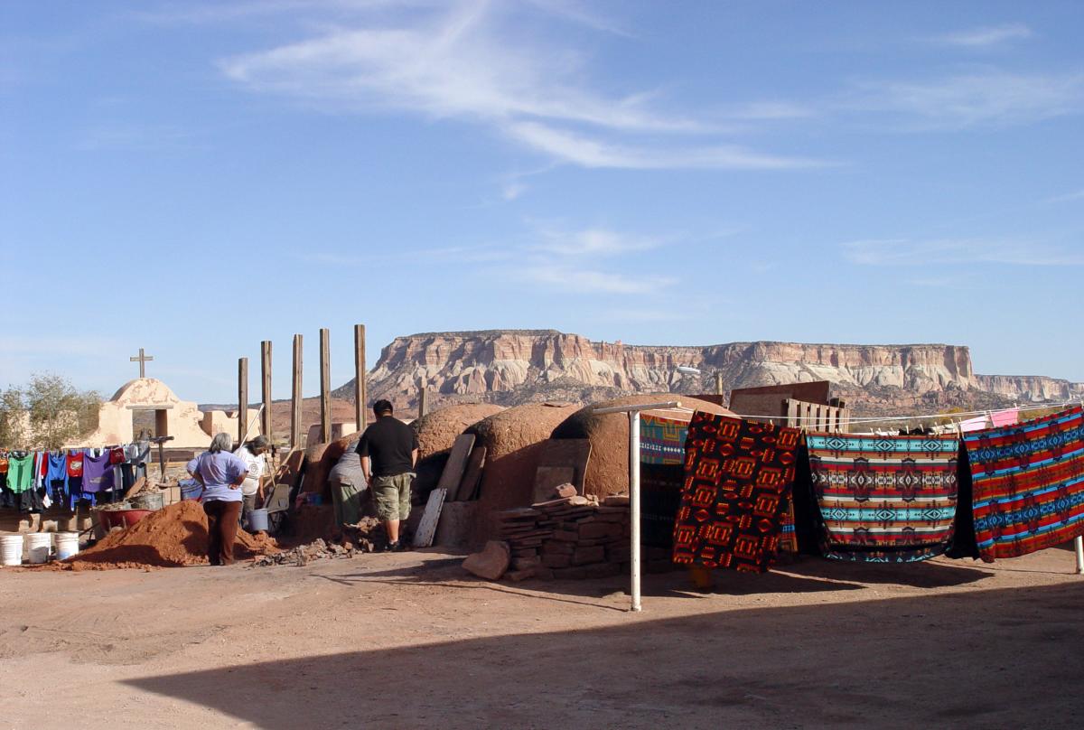 people standing next to hornos and traditional indigenous woven blankets, there is a church in the background and a large plateau