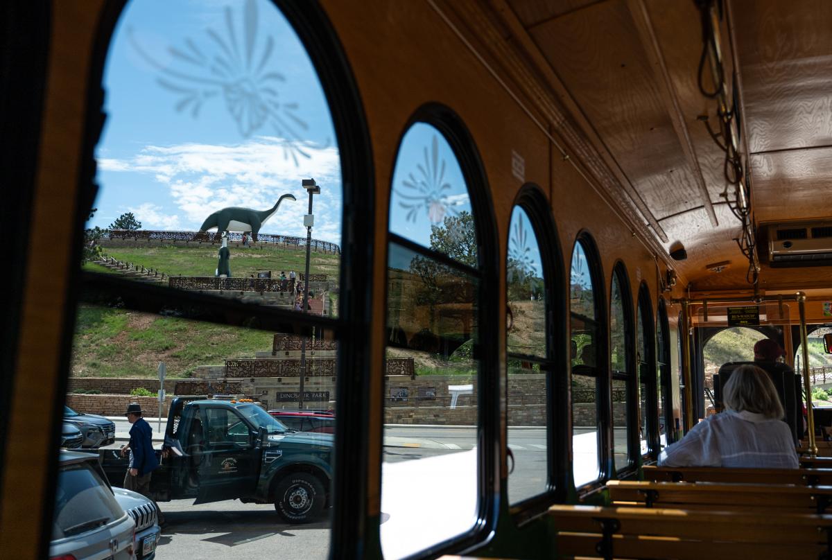View from inside the City View Trolley near Dinosaur Park, with the park’s hillside and large dinosaur sculpture visible through the arched windows. The trolley’s wooden interior, rows of windows, and seated passengers are visible on the right.