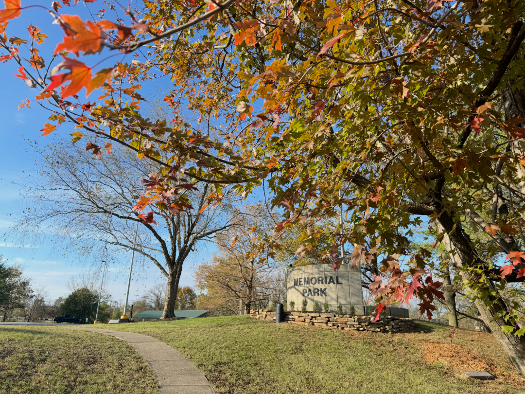 A park scene under a clear blue sky with vibrant autumn foliage. A pathway leads into the park, bordered by trees with red, orange, and yellow leaves. A stone sign in the background reads 'MEMORIAL PARK.' The ground is grassy with scattered fallen leaves.