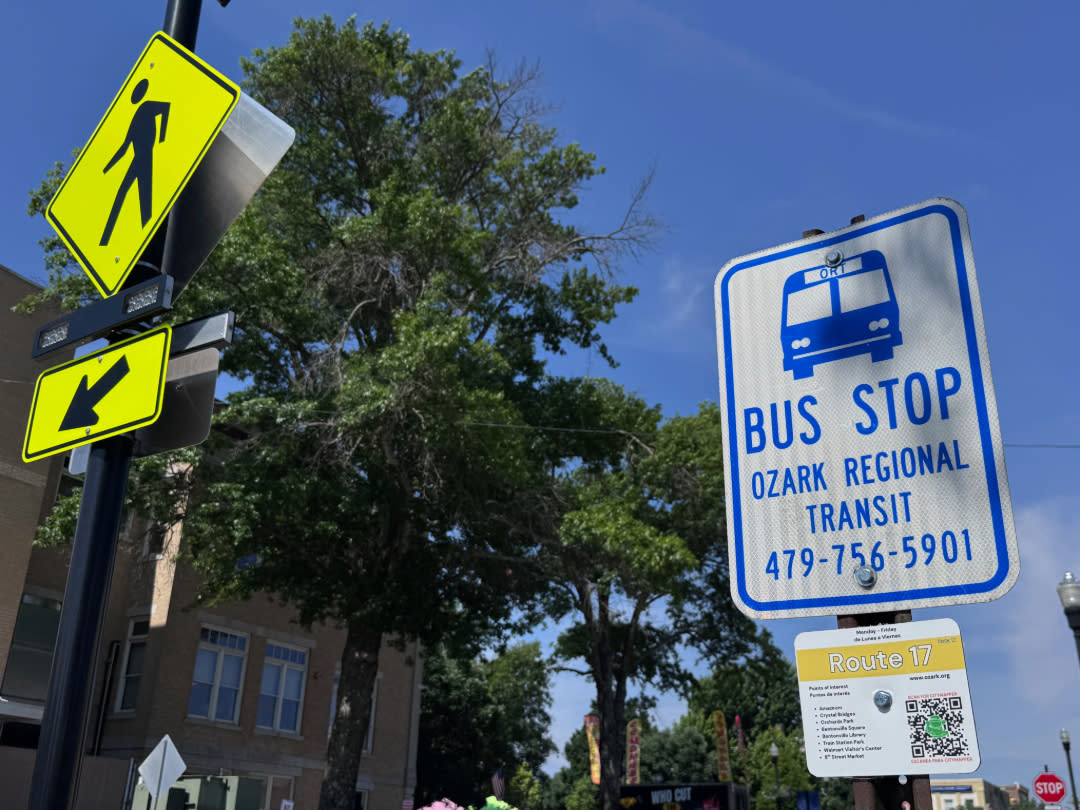 Bus stop sign for Ozark Regional Transit Route 17 beside a pedestrian crossing sign under a bright blue sky with trees and downtown buildings in the background.